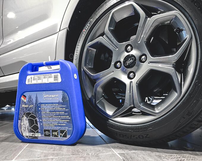 Zu sehen ist eine Packung Schneeketten neben einem montierten Reifen ohne Schneeketten